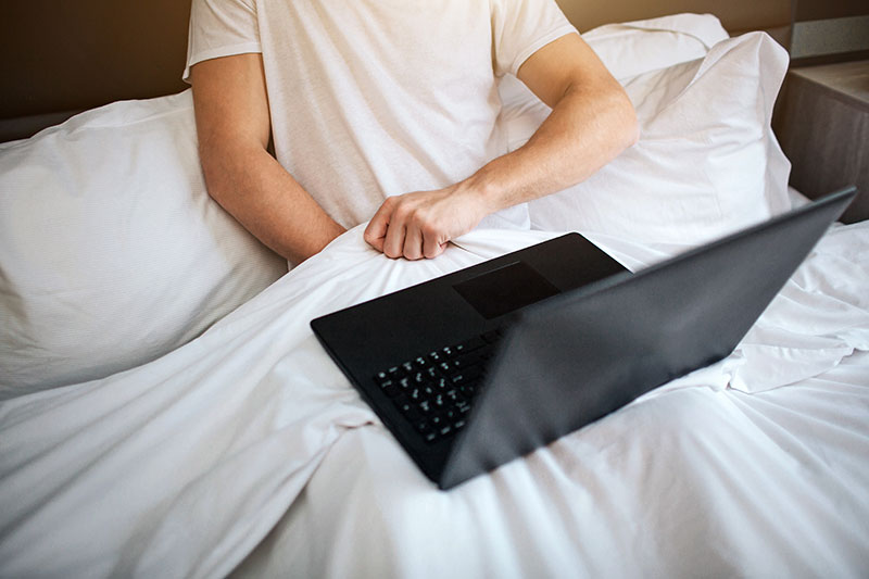 Mann liegt im Bett mit einem Laptop und greift unter die Decke