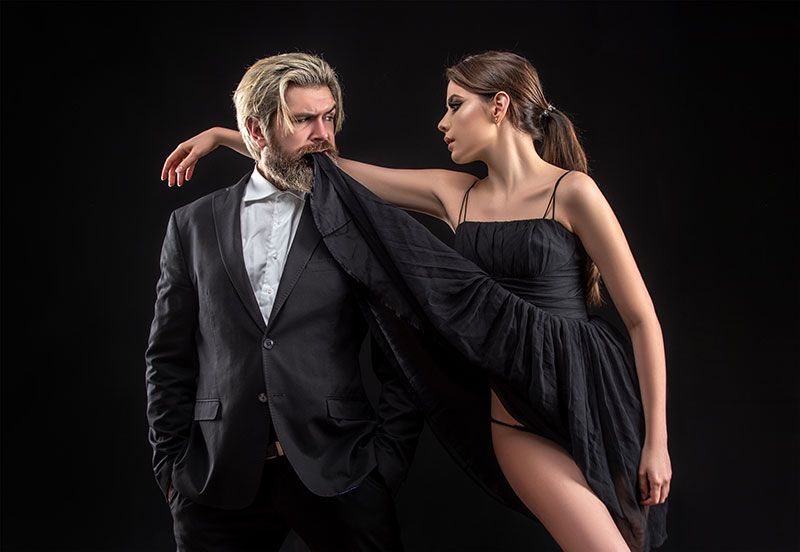 Mann mit Bart im Anzug nimmt schwarzes Kleid von einer Frau in den Mund