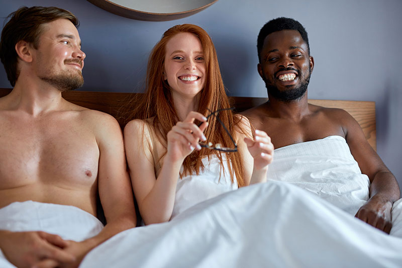 Rothaarige Frau liegt mit zwei Männern unter der Bettdecke