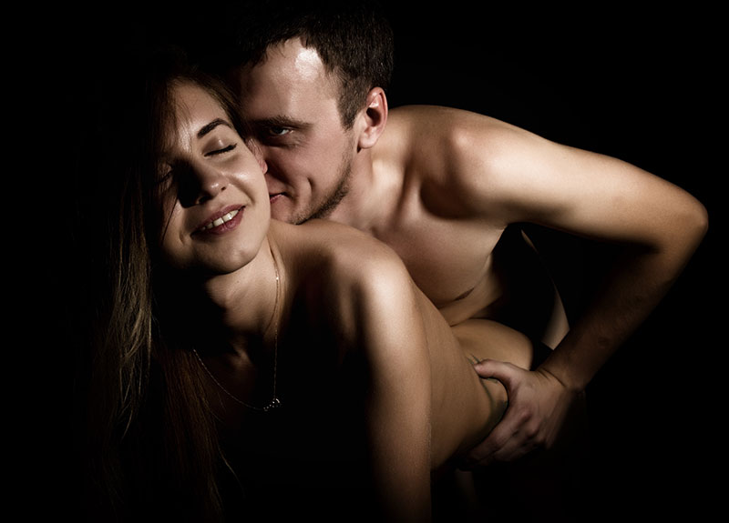 Nackter Mann hinter nackter Frau beim Sex vor einem schwarzen Hintergrund