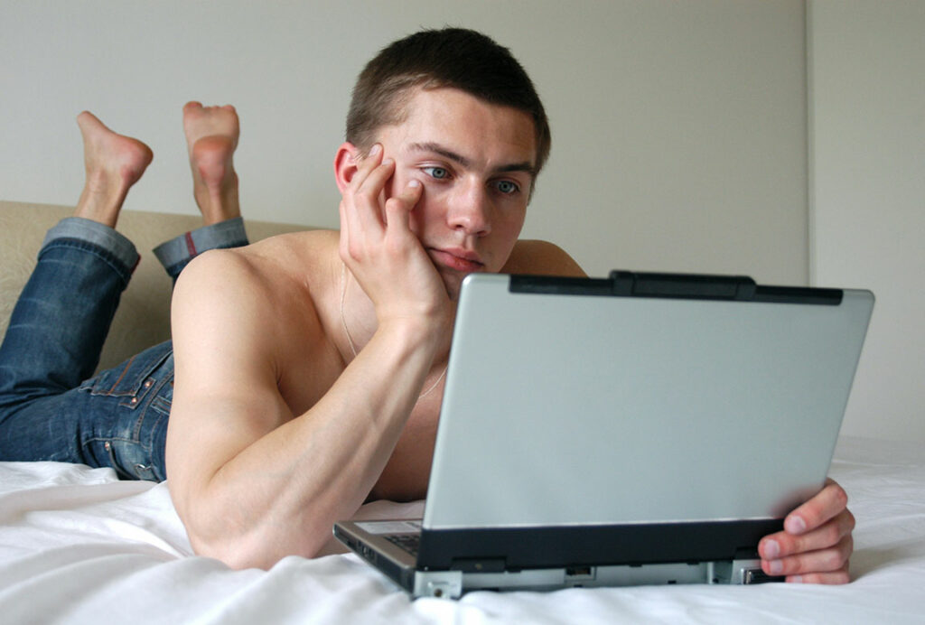Junger Mann liegt oberkörperfrei im Bett vor dem Laptop